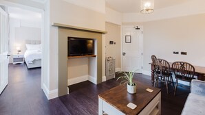 A modern hotel room with a bed, a TV mounted on a wooden shelf, a dining table with chairs, and a small potted plant. - Grace`s Guesthouse 68b (Tring)