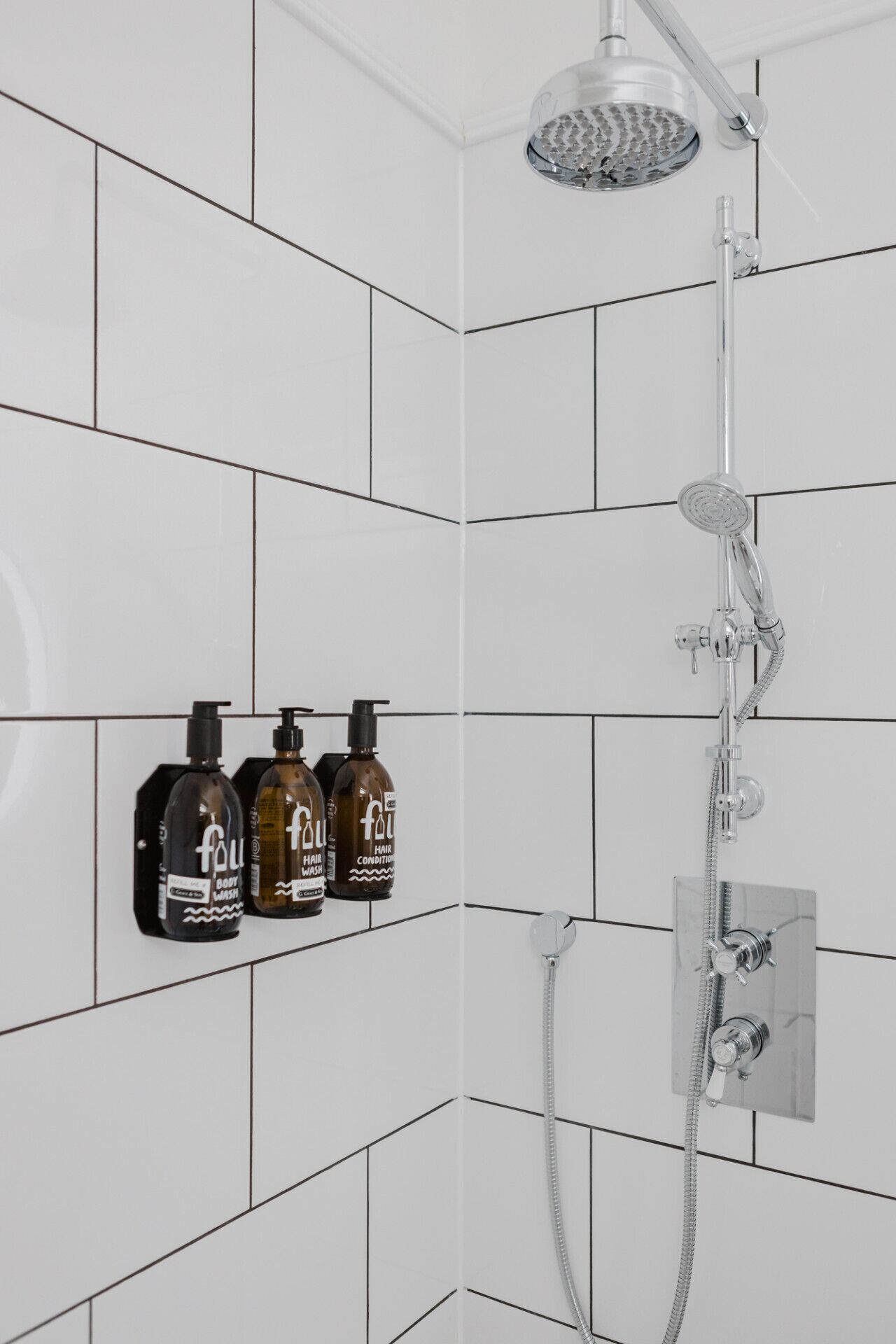 A shower with a handheld showerhead and a wall-mounted faucet. There are three bottles of hair care products on the wall.