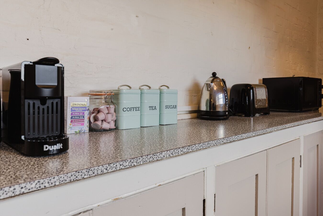 Un plan de travail de cuisine avec une machine à café Dualit, un bocal de sucre en morceaux rose et quatre récipients étiquetés pour le café, le thé et le sucre.