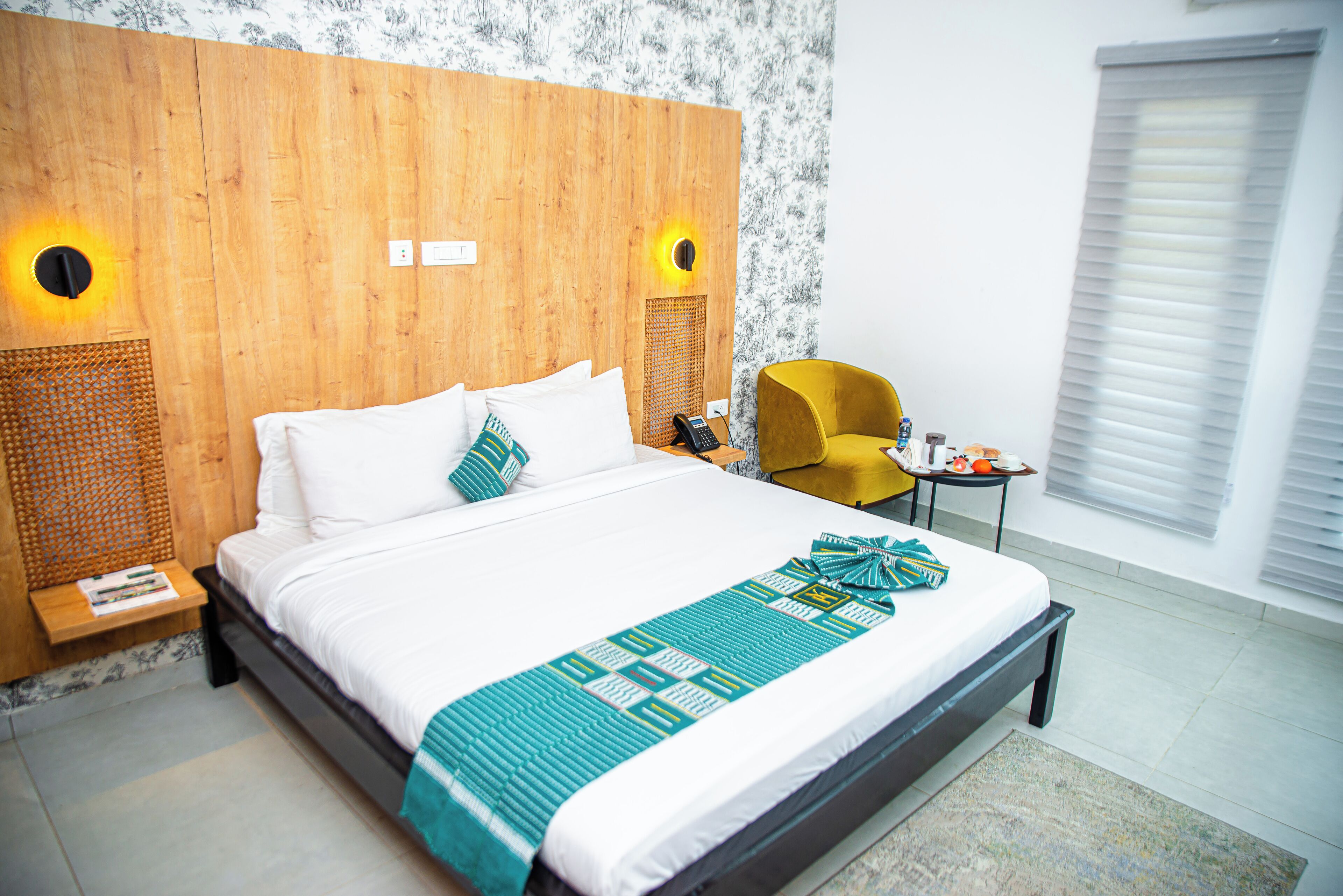 A hotel room with a large bed, wooden headboard, two bedside lamps, a small table with a phone, a yellow armchair, and a window with blinds.