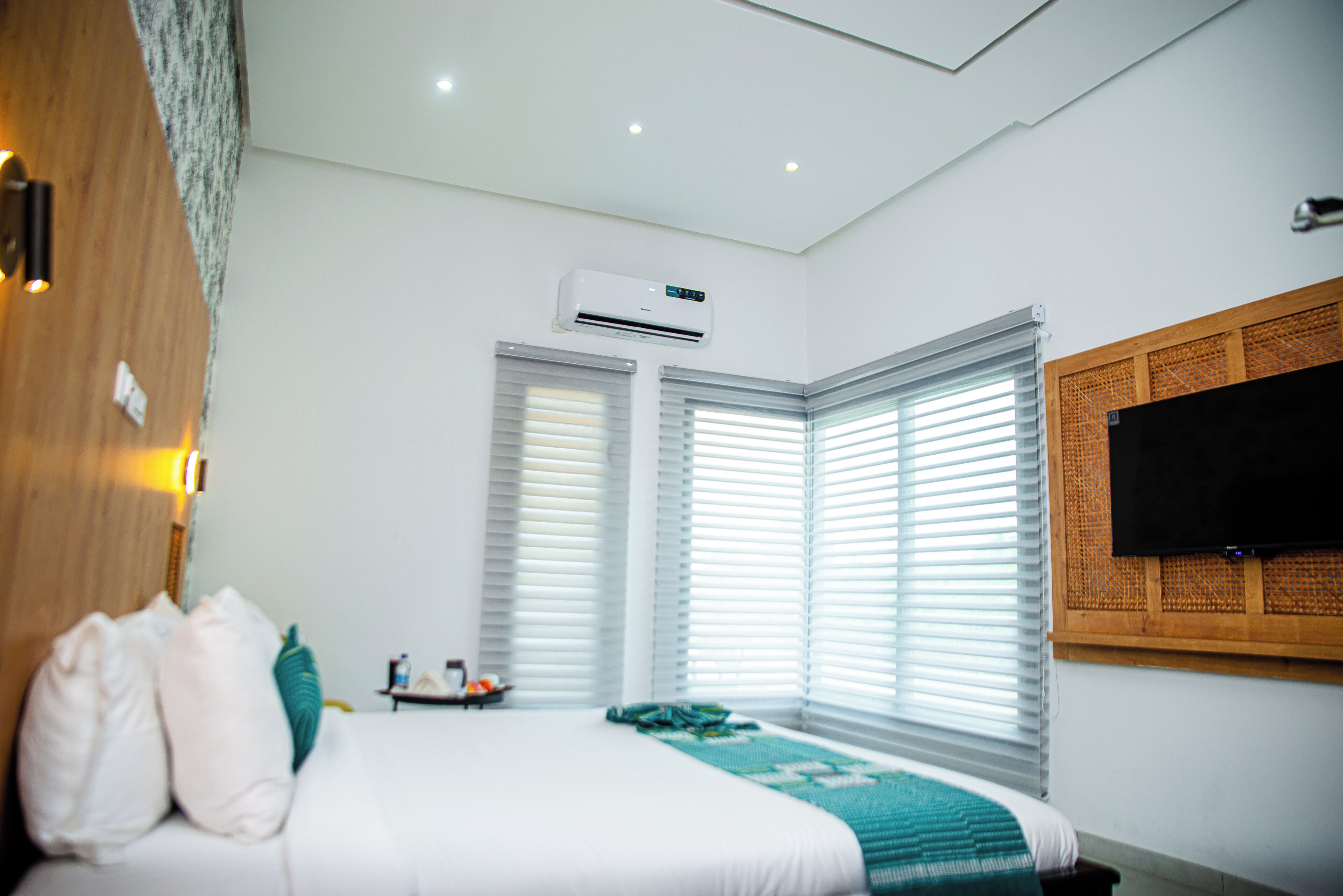 Ein Hotelzimmer mit Bett, Klimaanlage, Fenstern mit Jalousien, einem an der Wand montierten Fernseher und einem kleinen Tisch mit Gegenständen darauf.