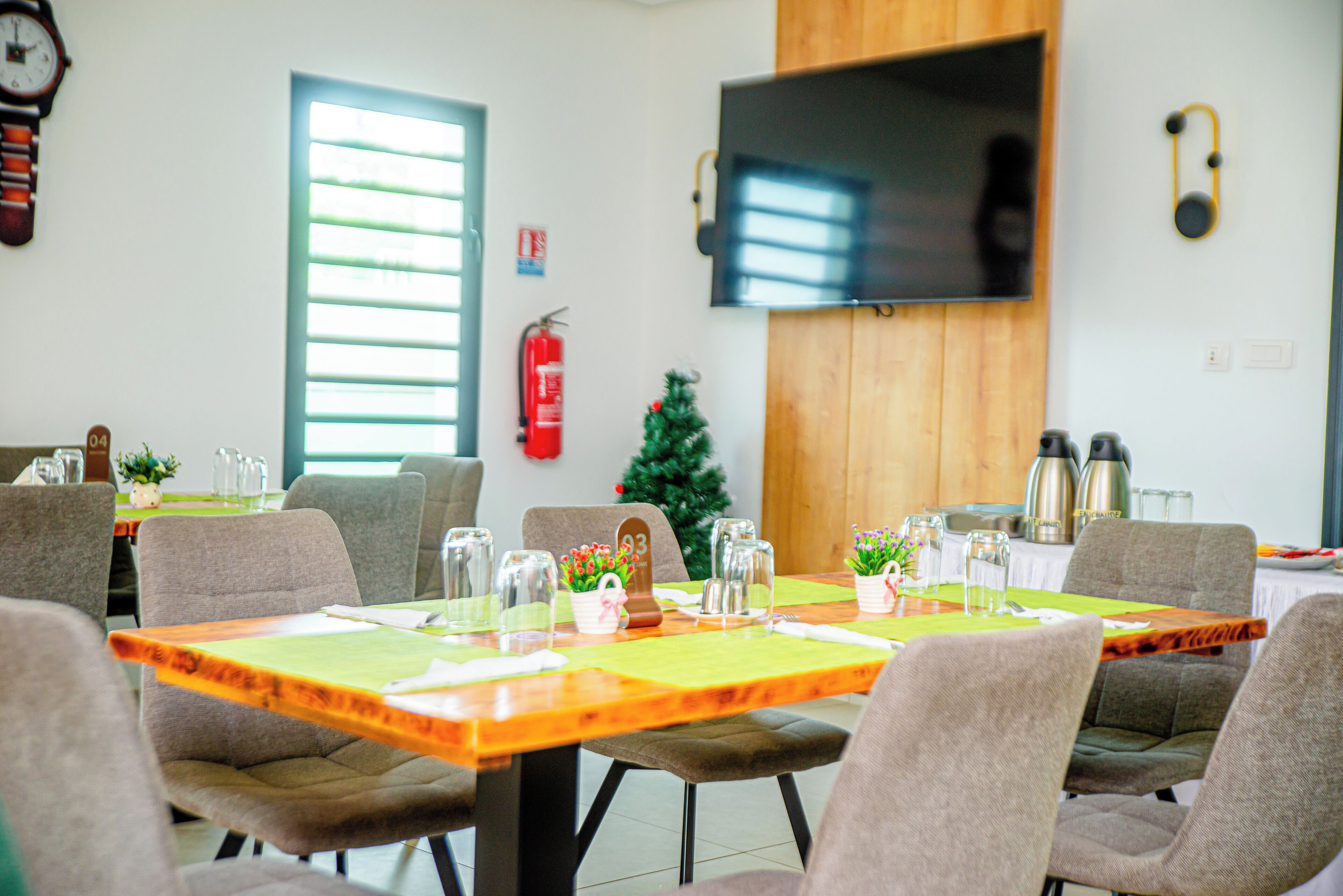 A dining area with tables set for four, chairs, a Christmas tree, a fire extinguisher, and a television mounted on the wall.