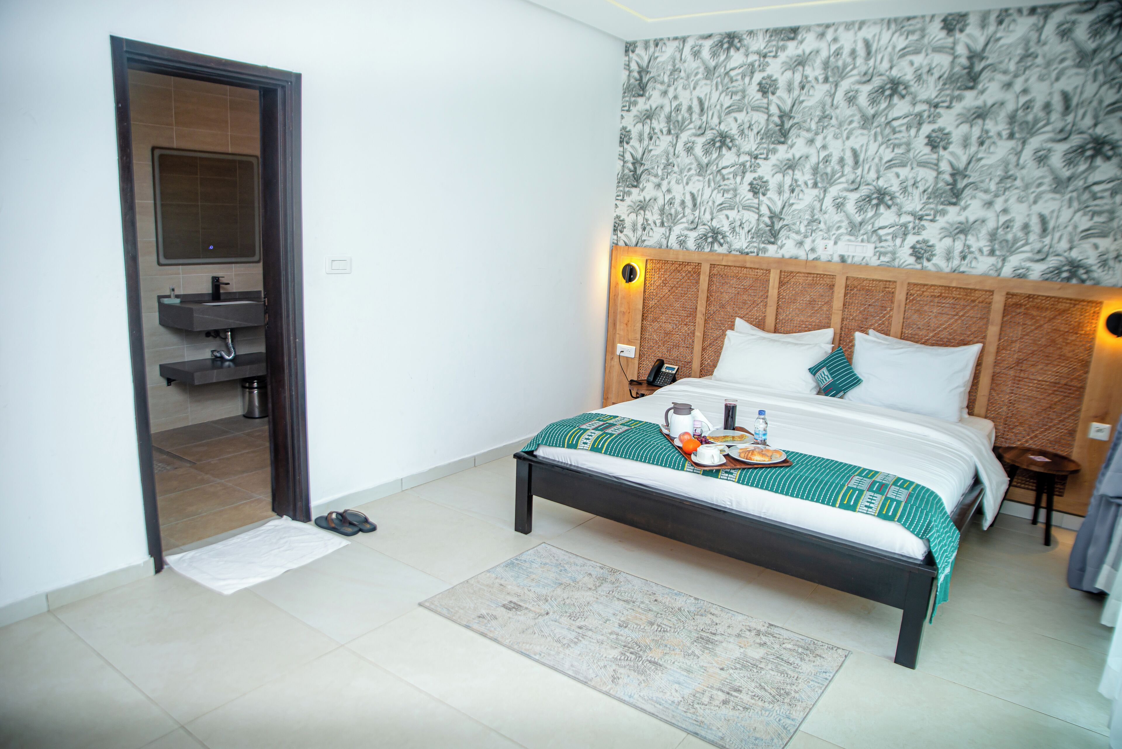 A hotel room with a bed, a bedside table, a nightstand, a mirror, and a bathroom with a sink and toilet.