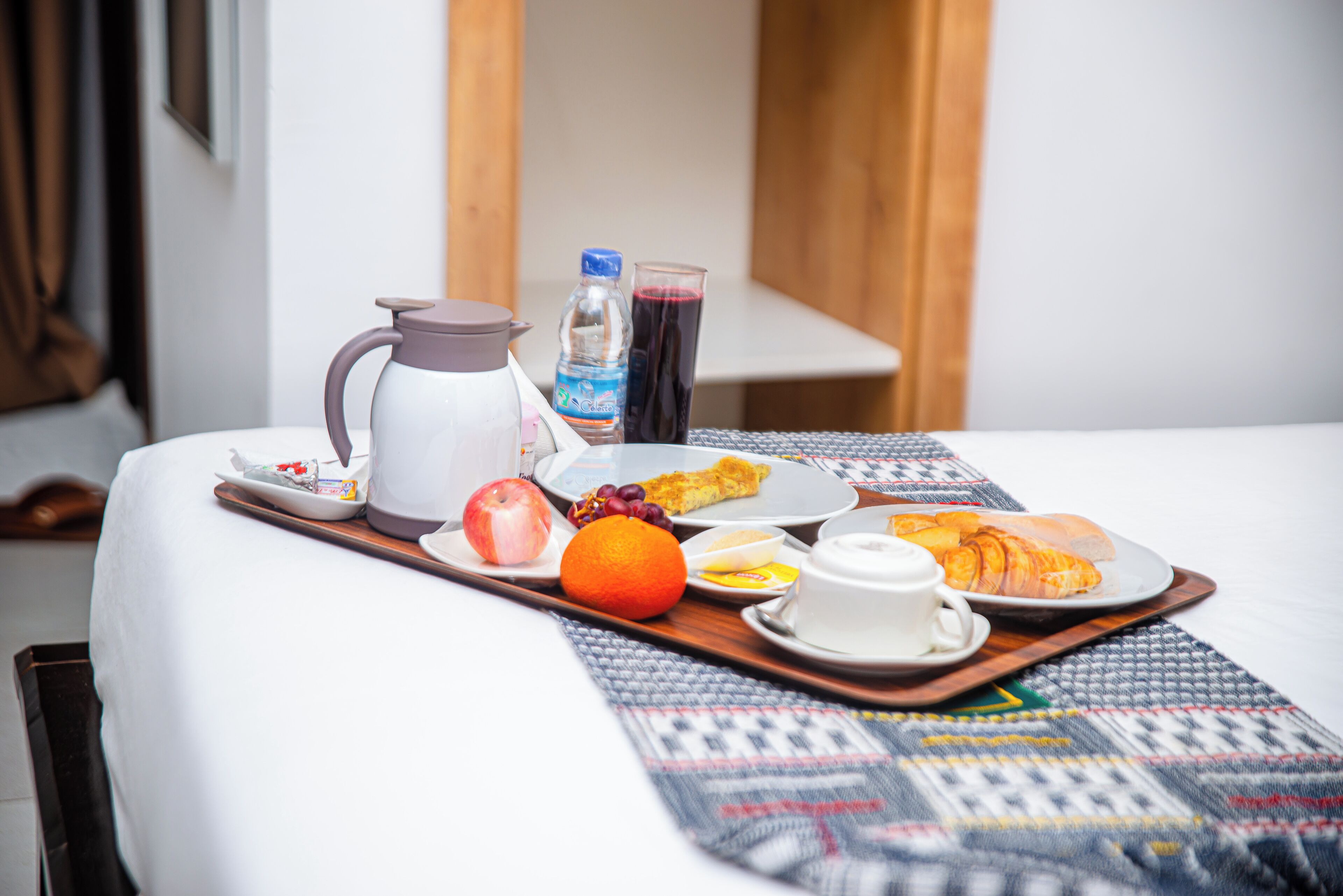 Una bandeja de desayuno ordenada sobre una cama, con jarra, botella de agua, jugo, fruta y un croissant.