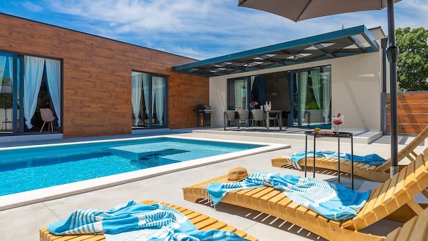 A modern outdoor pool area with wooden loungers, a covered seating area, and a glass-enclosed patio. - Lovely home in Rovinjsko selo with sauna (Rovinjsko selo)