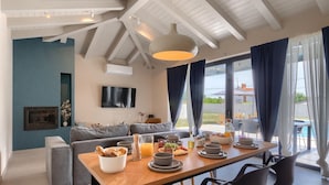 A modern living room with a wooden dining table set for breakfast, a grey sofa, and a TV mounted on the wall. - Lovely home in Rovinjsko selo with sauna (Rovinjsko selo)