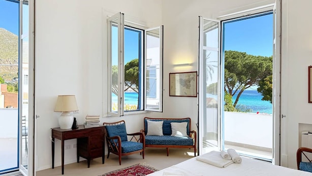 A bedroom with a bed, a desk, a chair, a rug, and large windows with a view of the sea.