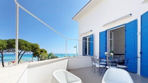 A balcony with a white chair, a table with plates, and blue shutters on a building overlooking a beach.