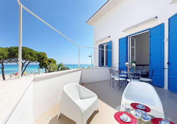 A balcony with a white chair, a table with plates, and blue shutters on a building overlooking a beach. - Solemar Sicilia - Villa Regina Elena (Palermo)