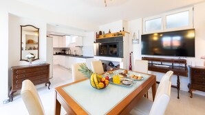 A modern kitchen with white cabinets, a dining table with fruit and pastries, and a large flat-screen TV.