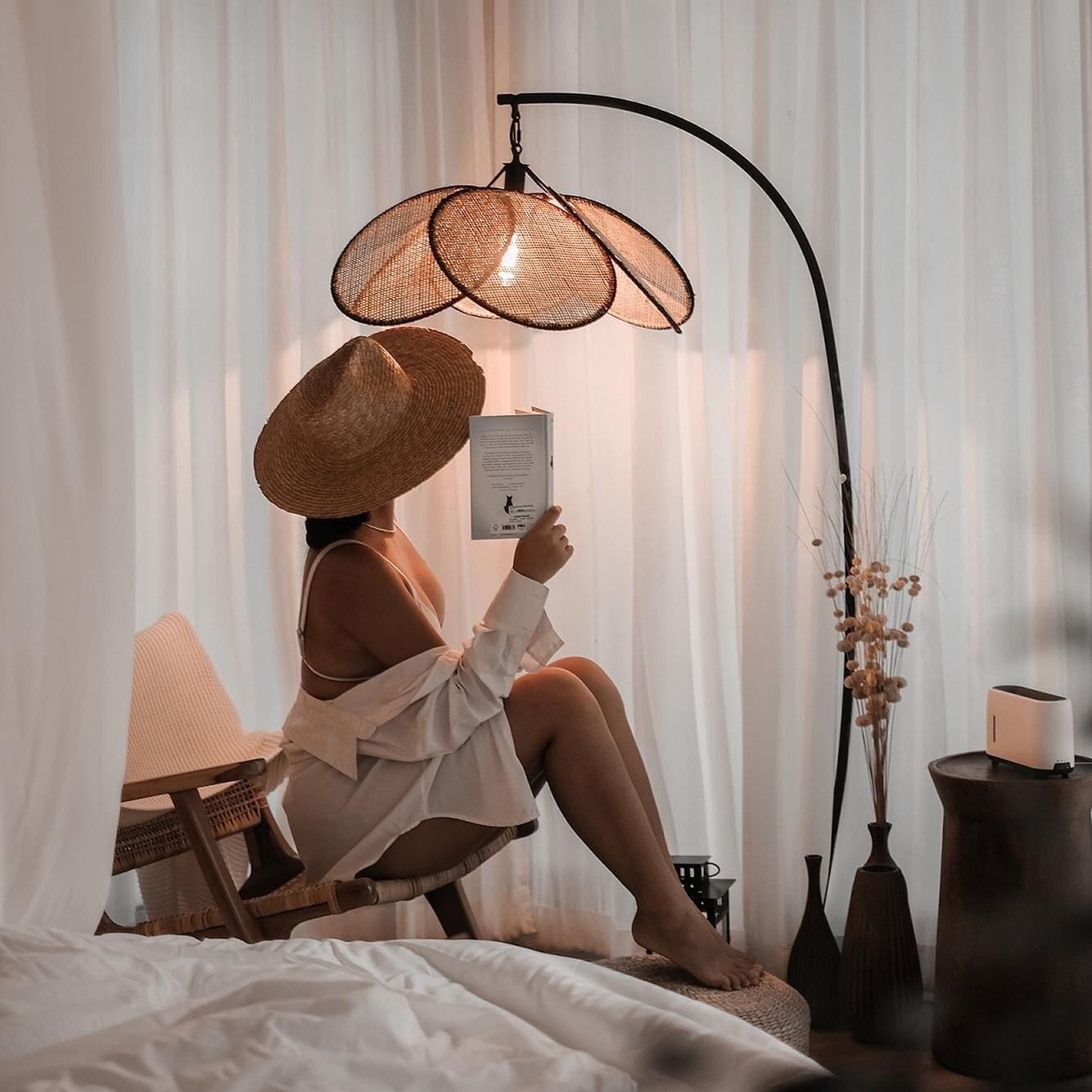 A person sitting on a bed reading a book, with a woven lamp and vases on a side table.