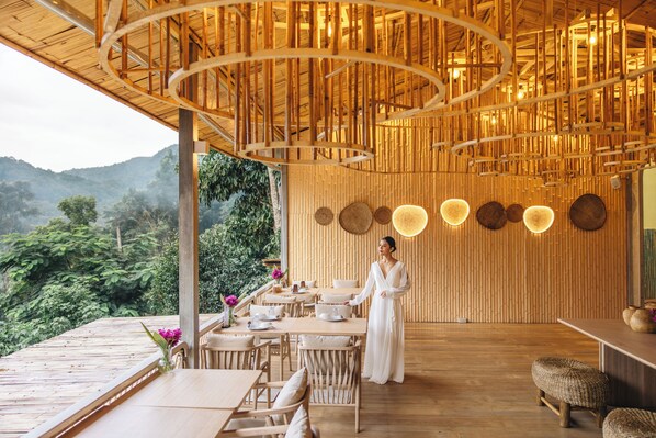 A wooden interior with a dining area, a person in a white dress, and a view of greenery outside. - Lannawild Chiang Mai (Mae On)