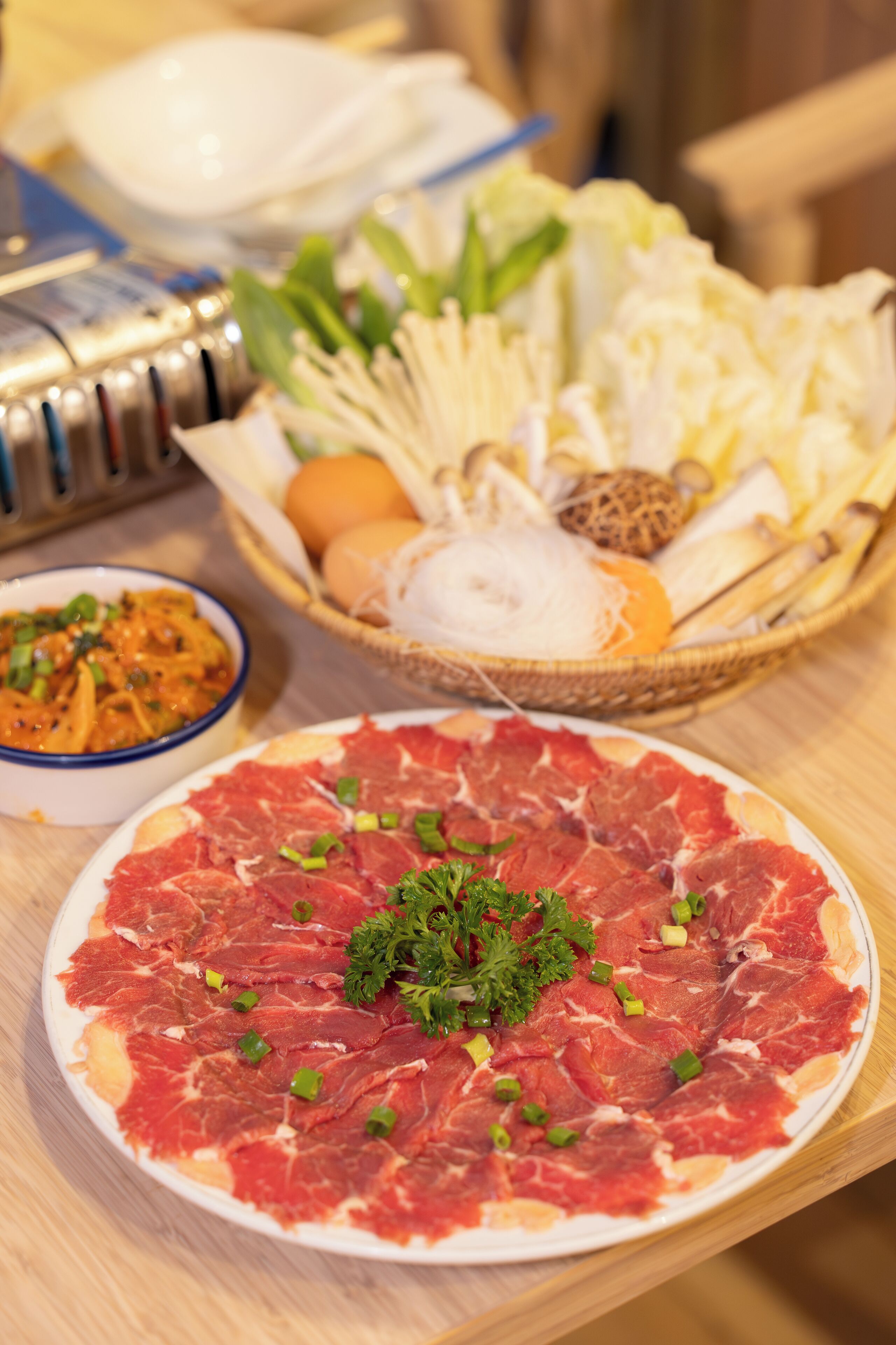 Une table à manger avec une assiette de viande crue, un panier de légumes et un bol de sauce.