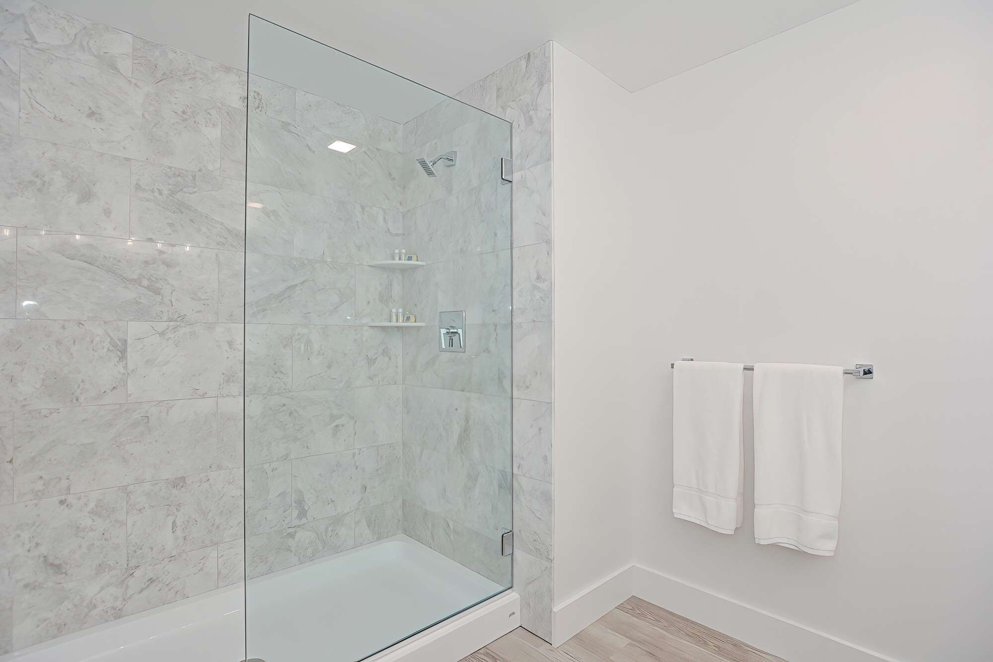 A modern bathroom with a glass-enclosed shower, white marble tiles, and a towel rack with two towels.