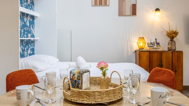 Ein Hotelzimmer mit einem Bett, einem Esstisch mit Stühlen, einem Korbwägelchen mit einer Rose und einer Wand mit Kunstwerken.
