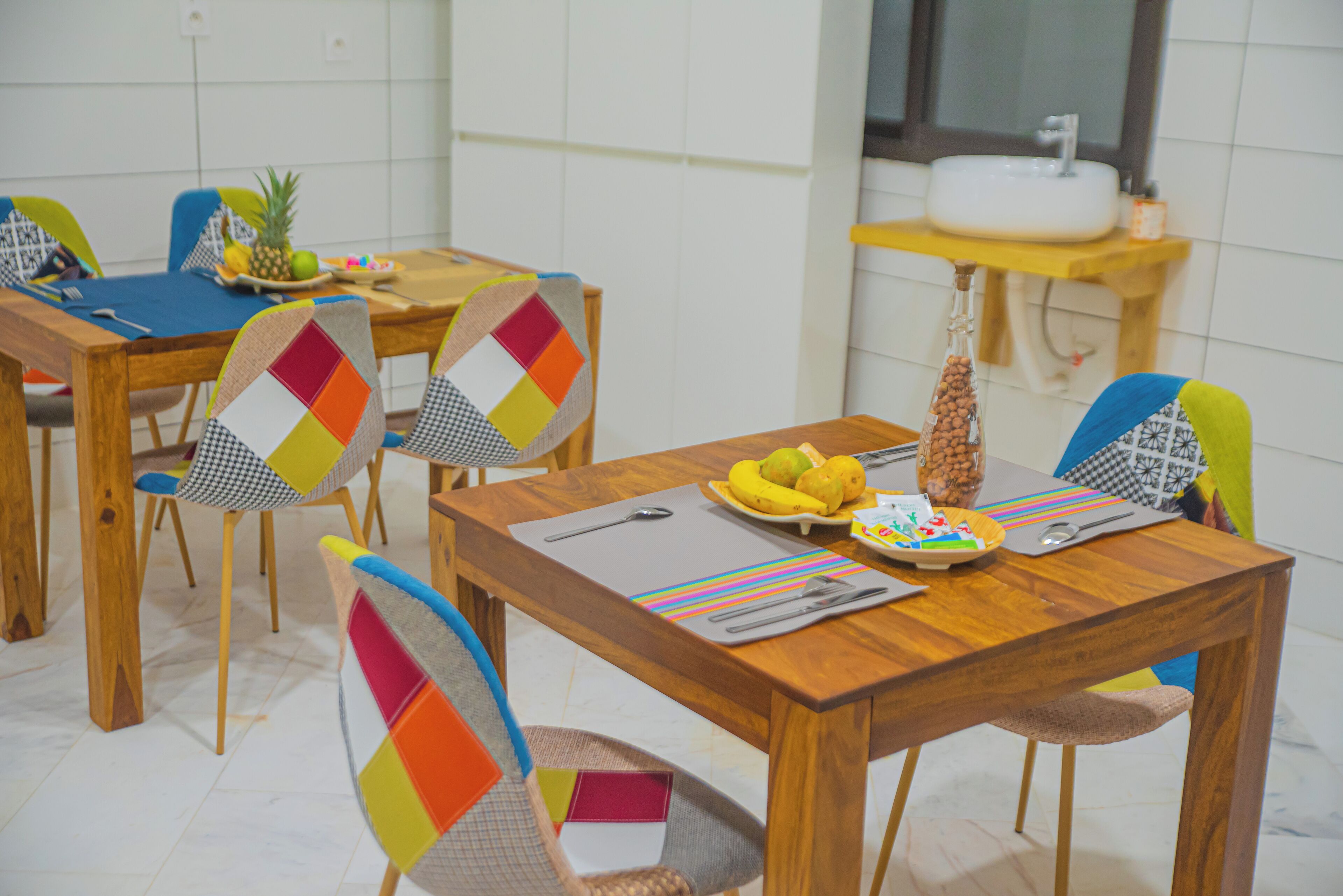 A dining area with wooden tables and chairs, a fruit bowl, and a sink in the background.