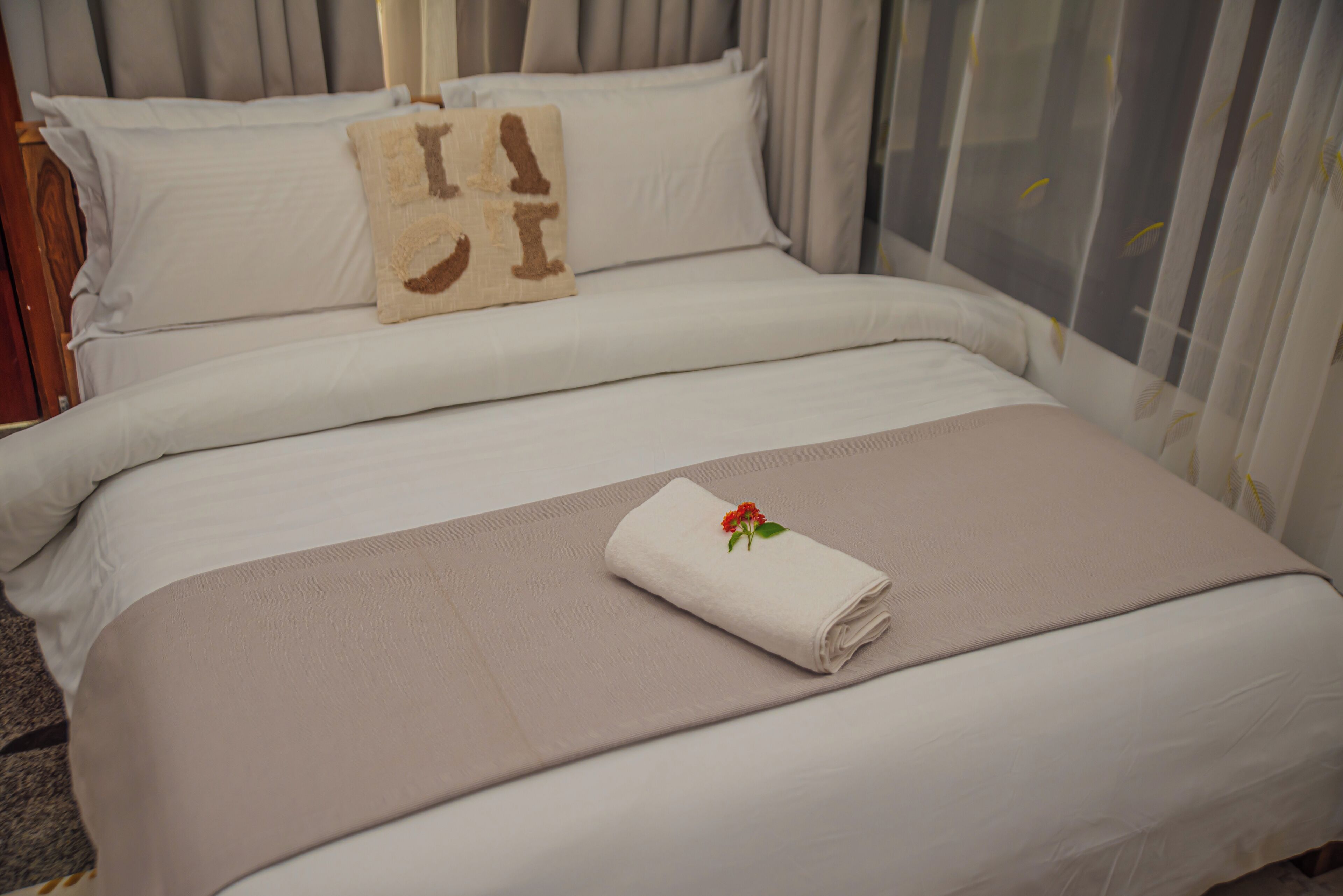 Ein ordentlich gemachtes Bett mit einer beigefarbenen Bettdecke, ein aufgerolltes weißes Handtuch mit roter Blume und ein dekoratives Kissen mit braunen und beigen Mustern.