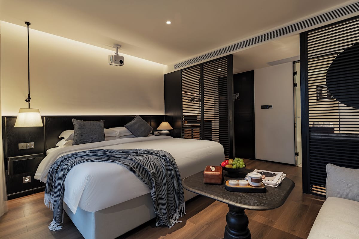 Ein modernes Hotelzimmer mit einem großen Bett, Nachttischen, einem kleinen Tisch mit Obstschale und Kerzen sowie einem Blick ins Freie durch ein Fenster mit Jalousien.