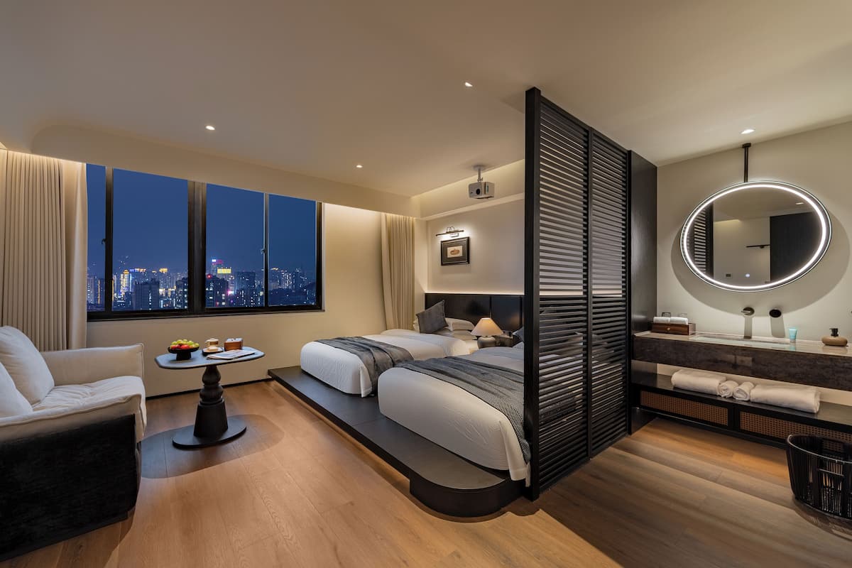 Ein modernes Hotelzimmer mit Blick auf die Stadt, einem Sofa, einem runden Spiegel, einem Schminktisch mit Waschbecken und einem Bett mit Nachttisch.