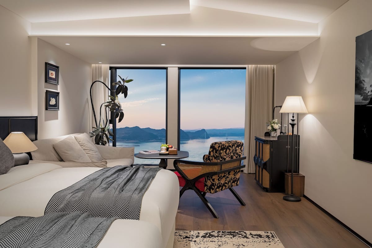 Ein Hotelzimmer mit einem großen Bett, einer Sitzecke mit Meerblick, einem kleinen Tisch mit einer Pflanze und einer Lampe.