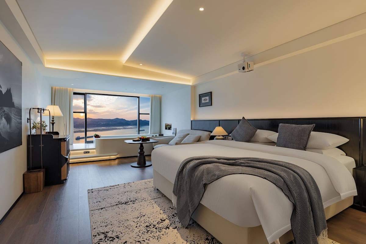Ein modernes Hotelzimmer mit einem großen Bett, Blick auf den Sonnenuntergang durch das Fenster, einem kleinen Tisch mit Lampe und einem Nachttisch.