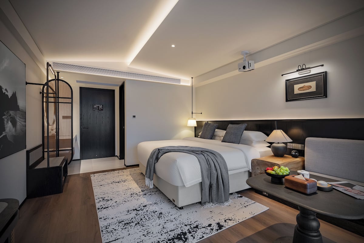Ein modernes Hotelzimmer mit Bett, Nachttisch, Lampe und einer kleinen Sitzecke mit Sofa und Tisch.