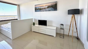 Ein modernes Schlafzimmer mit einem Bett, einem Fernseher auf einem weißen Schrank, einem Gemälde mit Strandmotiv und einer Stehlampe.