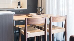 A modern kitchen with dark cabinetry, a white countertop, and a built-in coffee machine. There is a dining table with two chairs and a pitcher on it.
