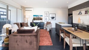 A modern living room with a sofa, coffee table, and a dining area with a wooden table and chairs. There is a kitchen area with a refrigerator and a sink.