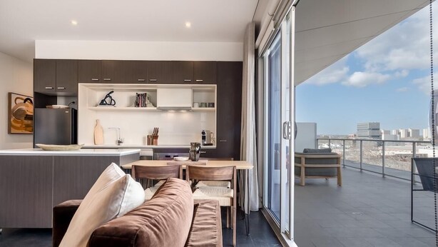A modern kitchen with dark cabinets, a dining area with a brown sofa, and a balcony with a city view.