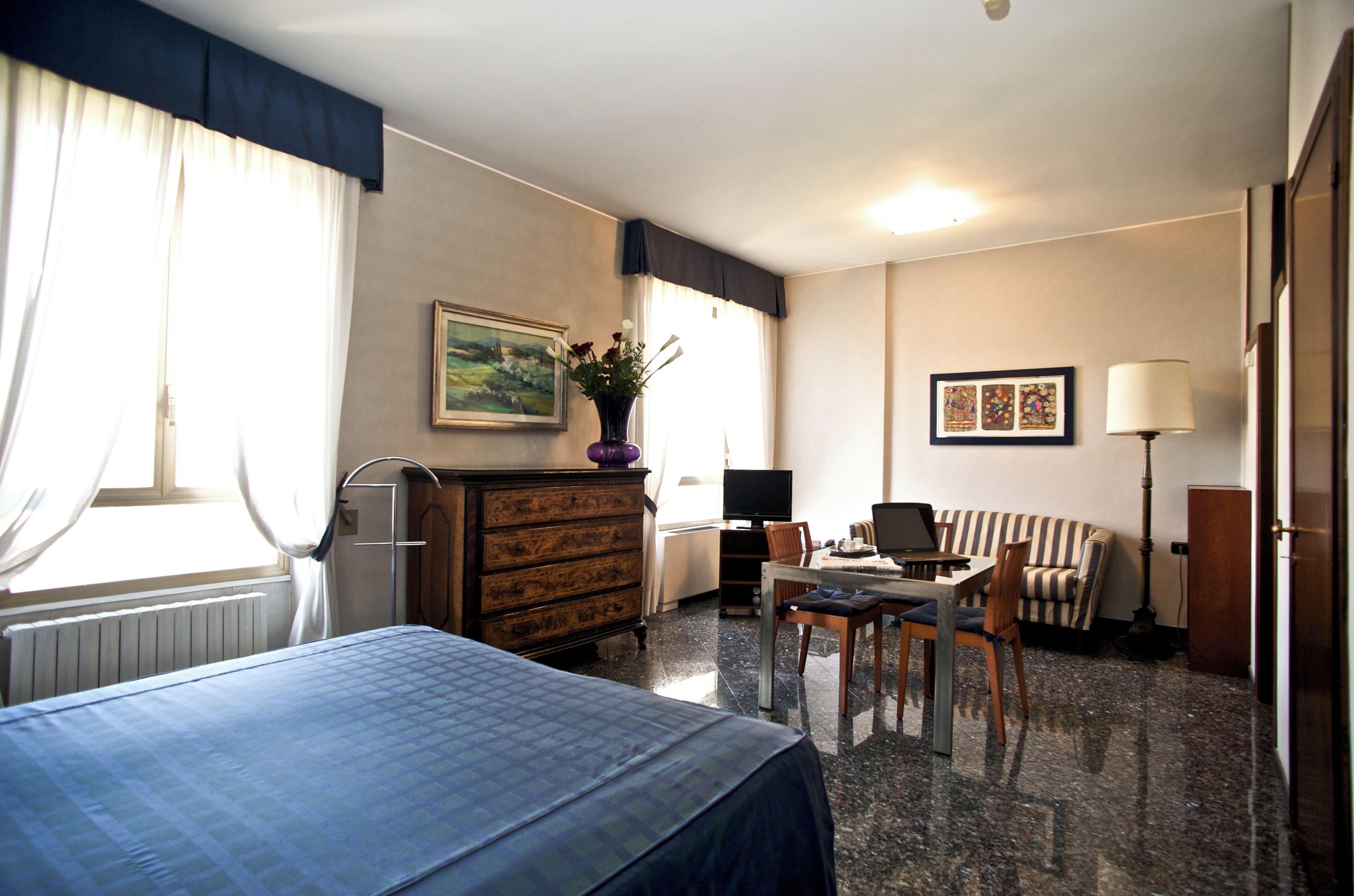 A hotel room with a bed, a desk with a chair, a lamp, a dresser, and a painting on the wall.