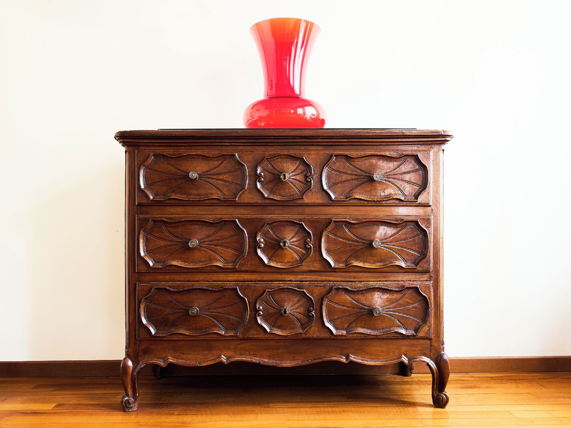 Une commode en bois avec six tiroirs, sur laquelle est posé un vase rouge.