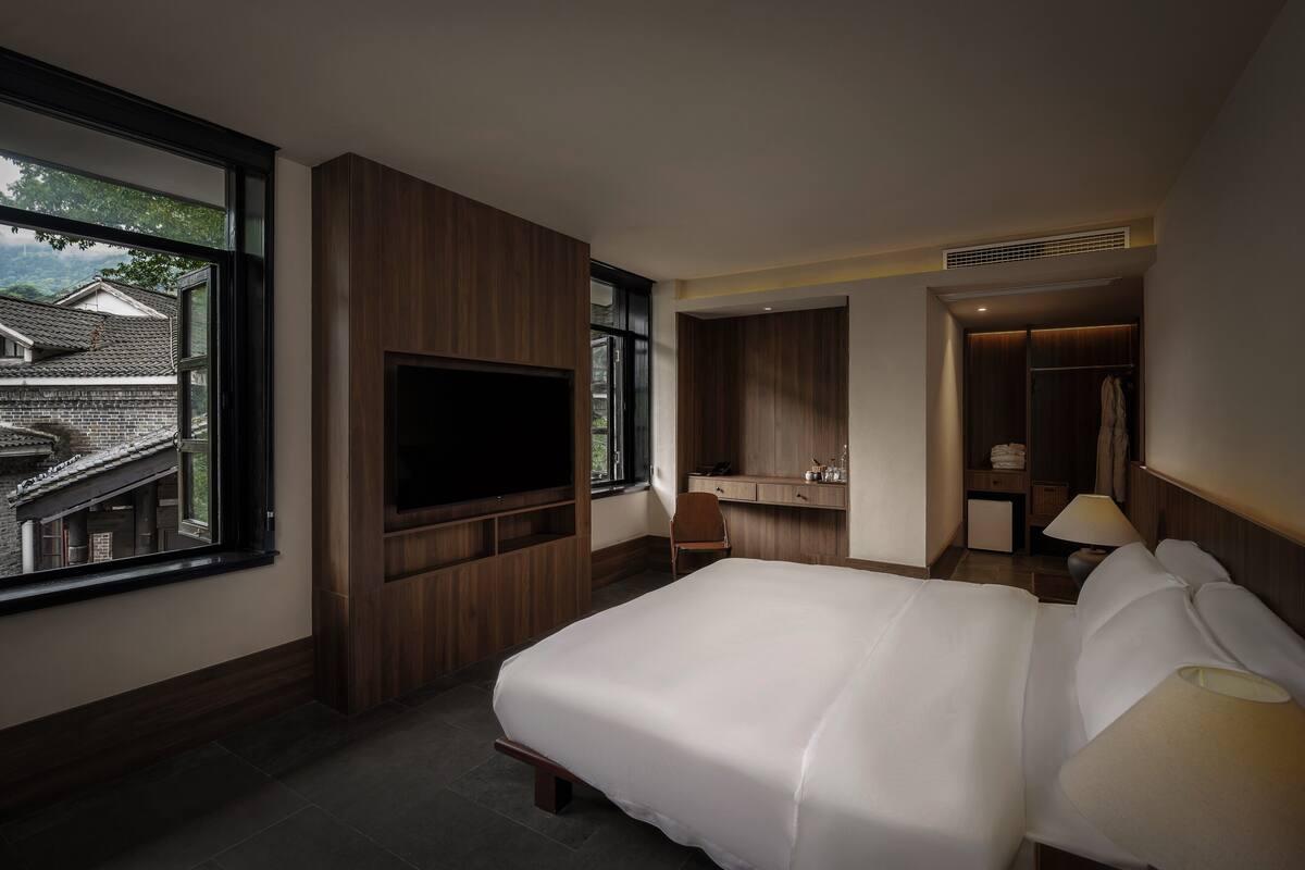 Ein Hotelzimmer mit einem großen Bett, einem an der Wand montierten Fernseher, einem Schreibtisch mit Stuhl und einem Badezimmer mit Spiegel und Handtuchhalter.
