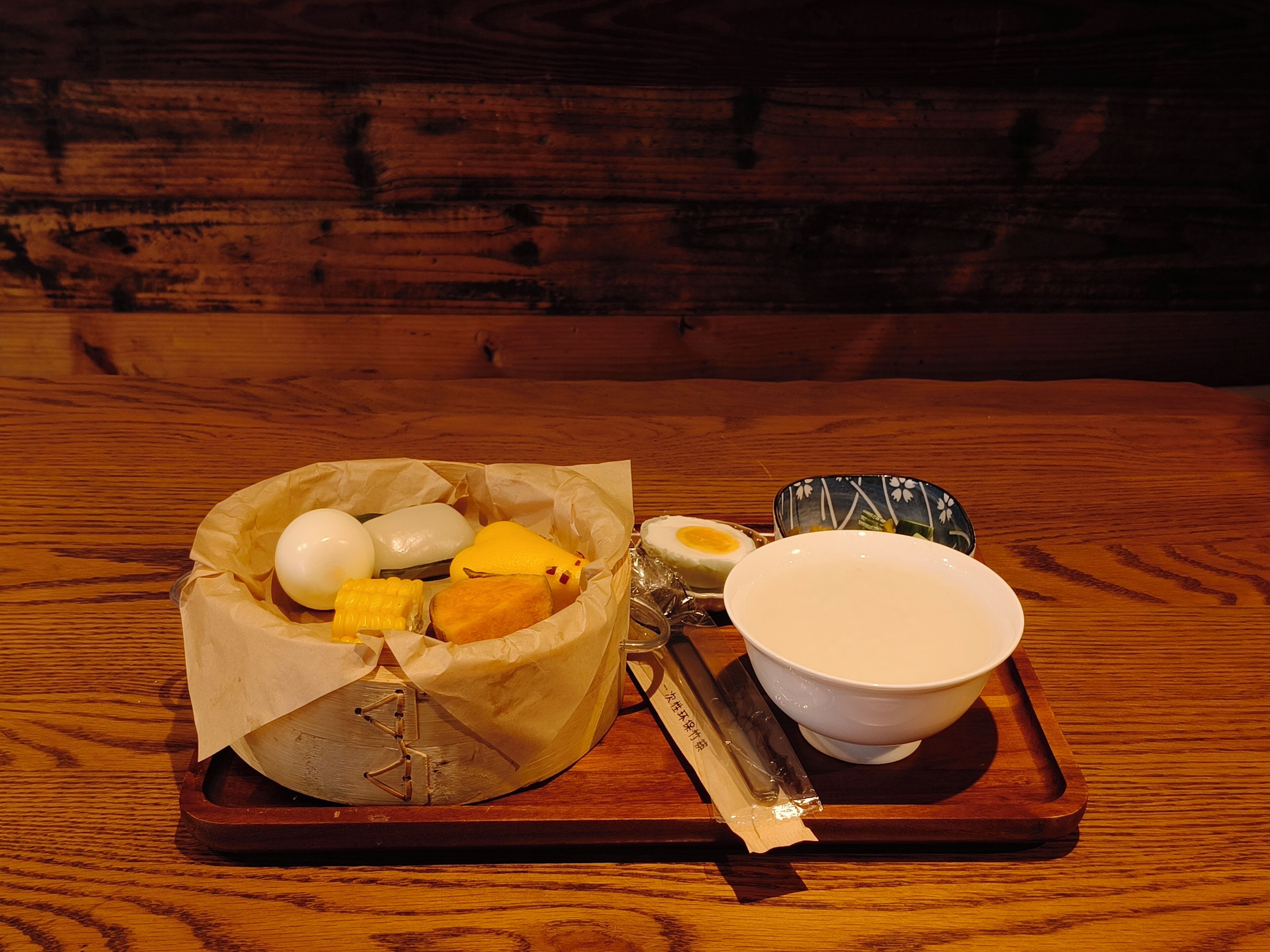 Ein Holztisch mit einer Tablettablage, auf der eine Schüssel mit Essen steht, darunter Eier und ein Stück Fleisch, sowie eine kleine Sojasaucen-Behälter.