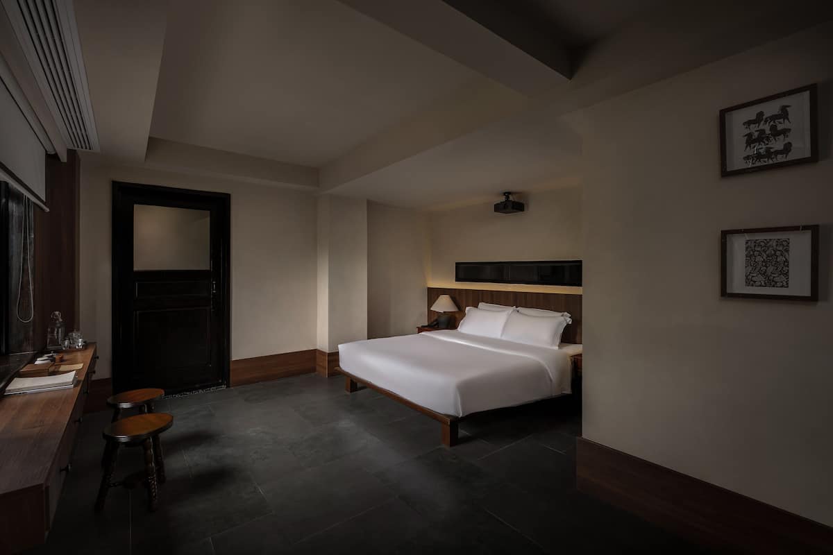Ein Hotelzimmer mit einem Bett, einem Schreibtisch, zwei Hocker, einer Tür und gerahmten Kunstwerken an der Wand.