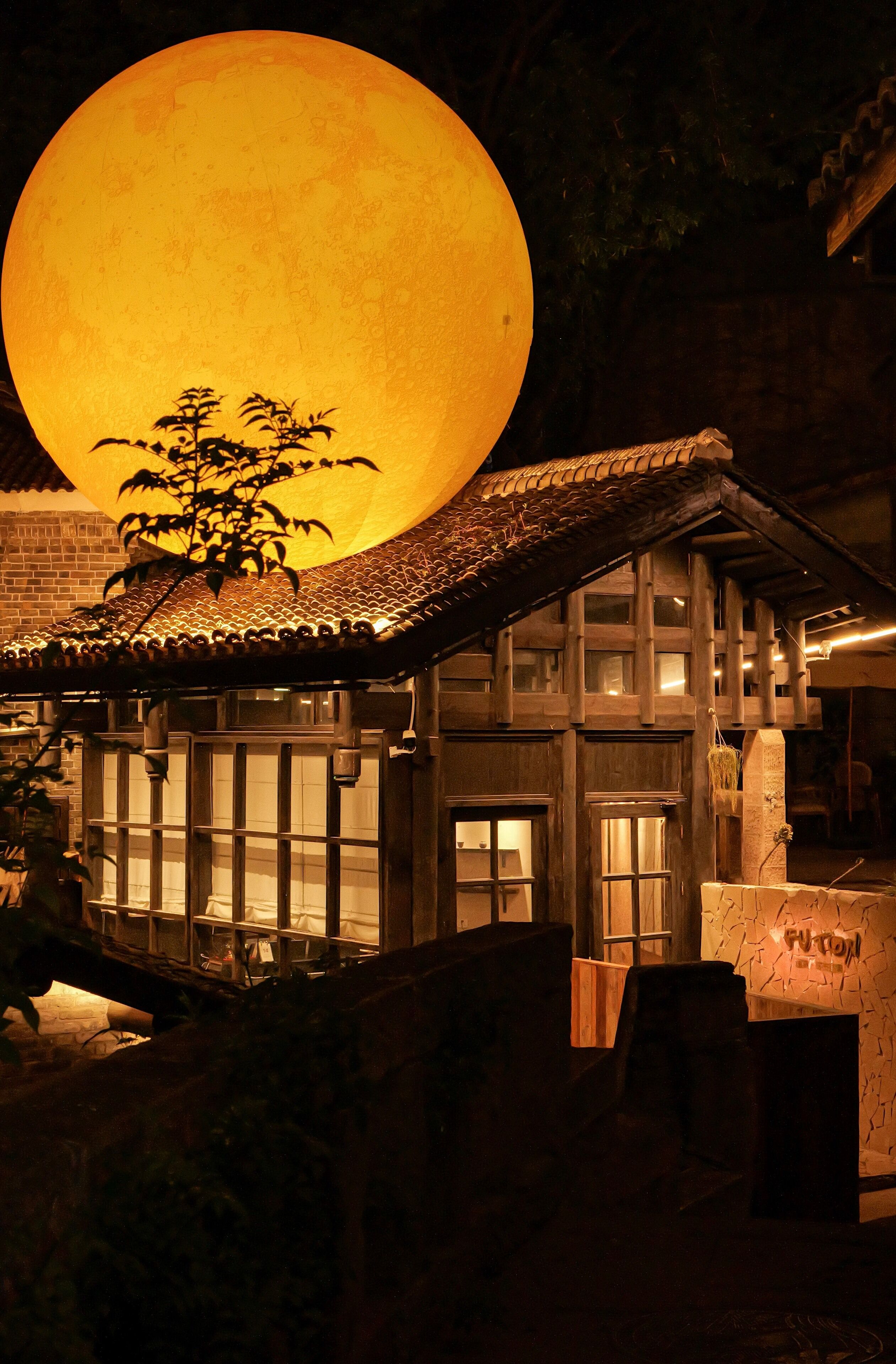 Un edificio tradicional con techo de tejas y paredes de madera, una luna grande al fondo y un pequeño árbol en primer plano.