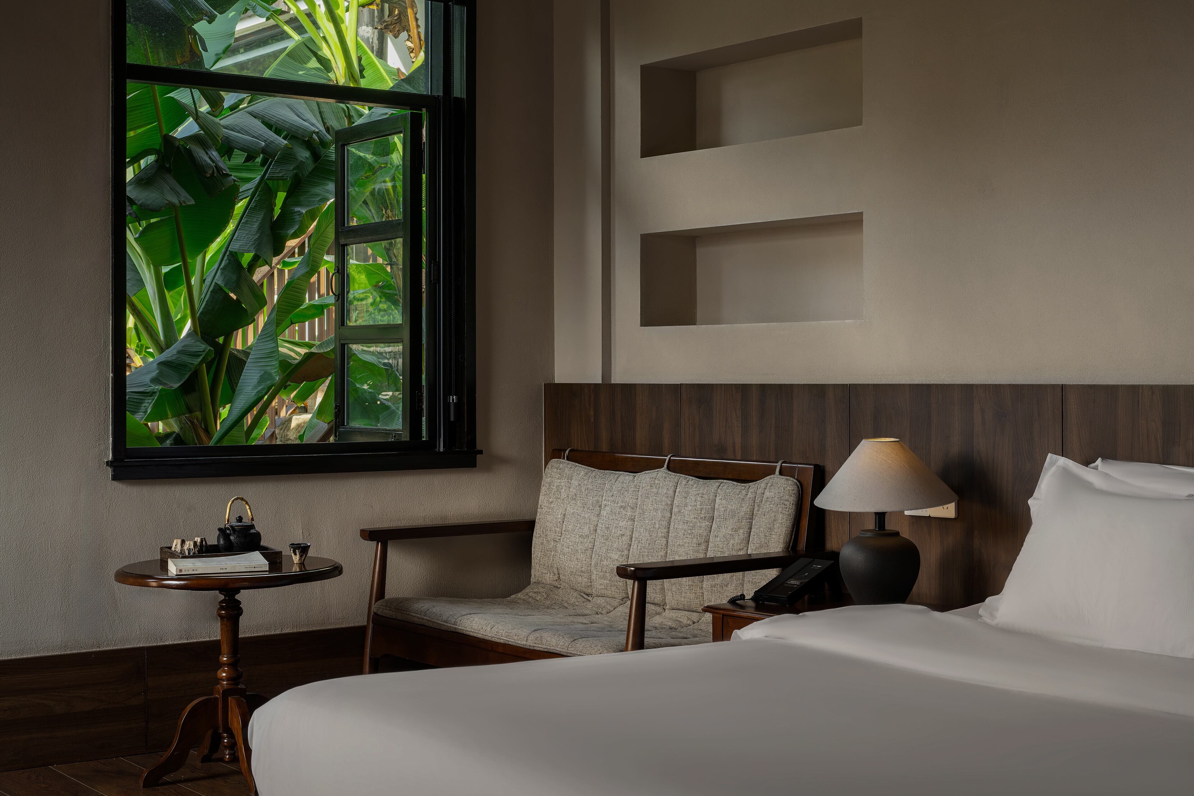 A hotel room with a bed, a chair, a small table with a lamp, and a window with a view of greenery.