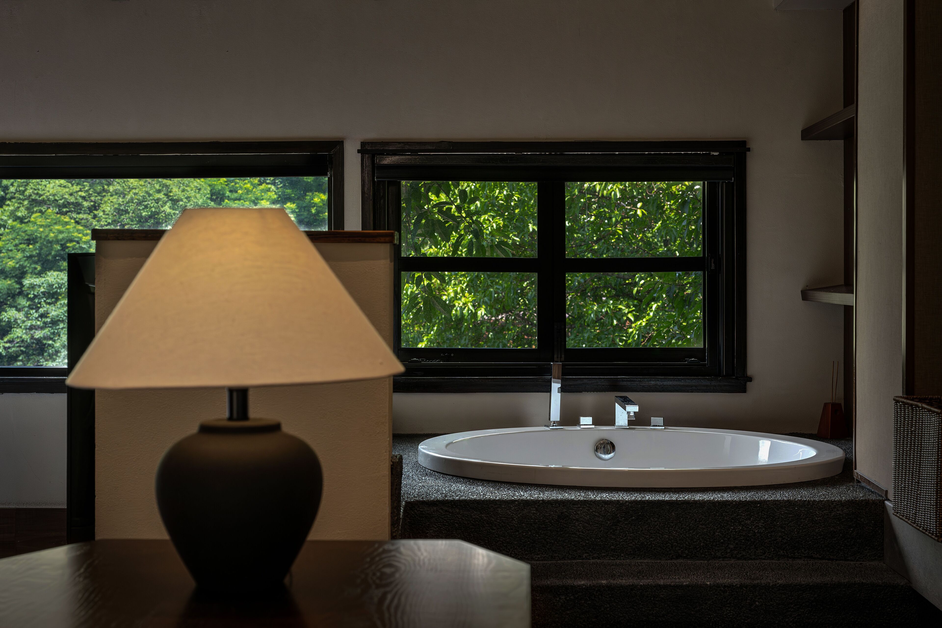 A bathroom with a bathtub, a window with a view of trees, and a lamp on a table.