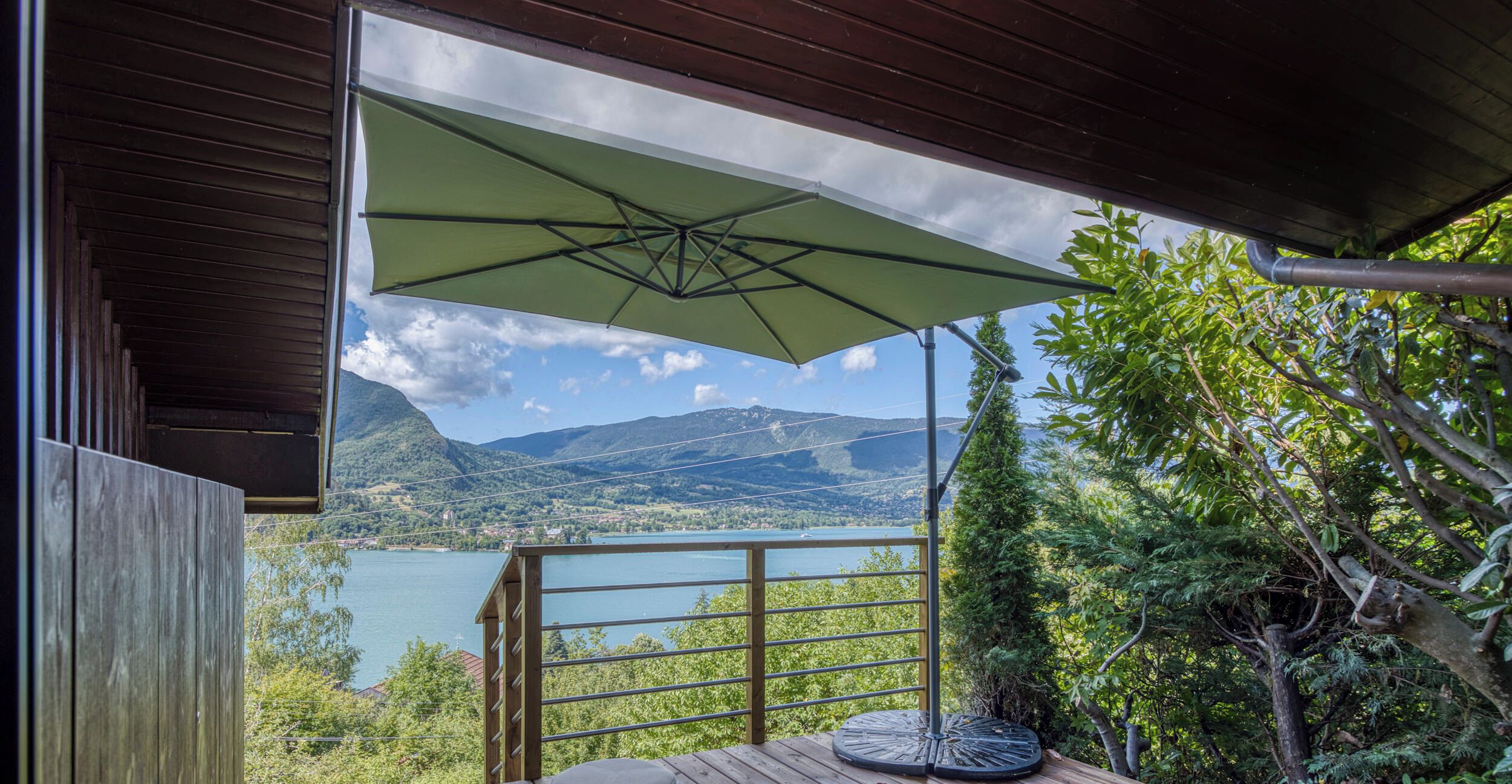 A wooden deck with a green umbrella, two stools, and a view of a lake and mountains.