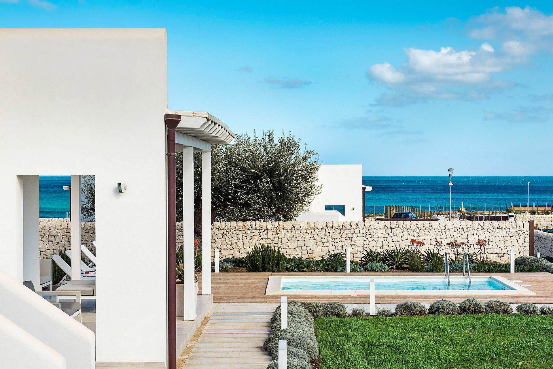 A modern outdoor pool area with a stone wall, a wooden deck, and a view of the sea.