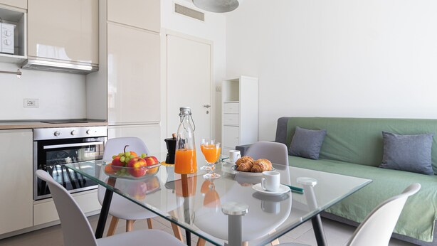 A modern kitchen with an oven, a glass table with chairs, a green sofa, and breakfast items like fruit, juice, and pastries. - Italianway Comfort - Watt 8 (Milan)