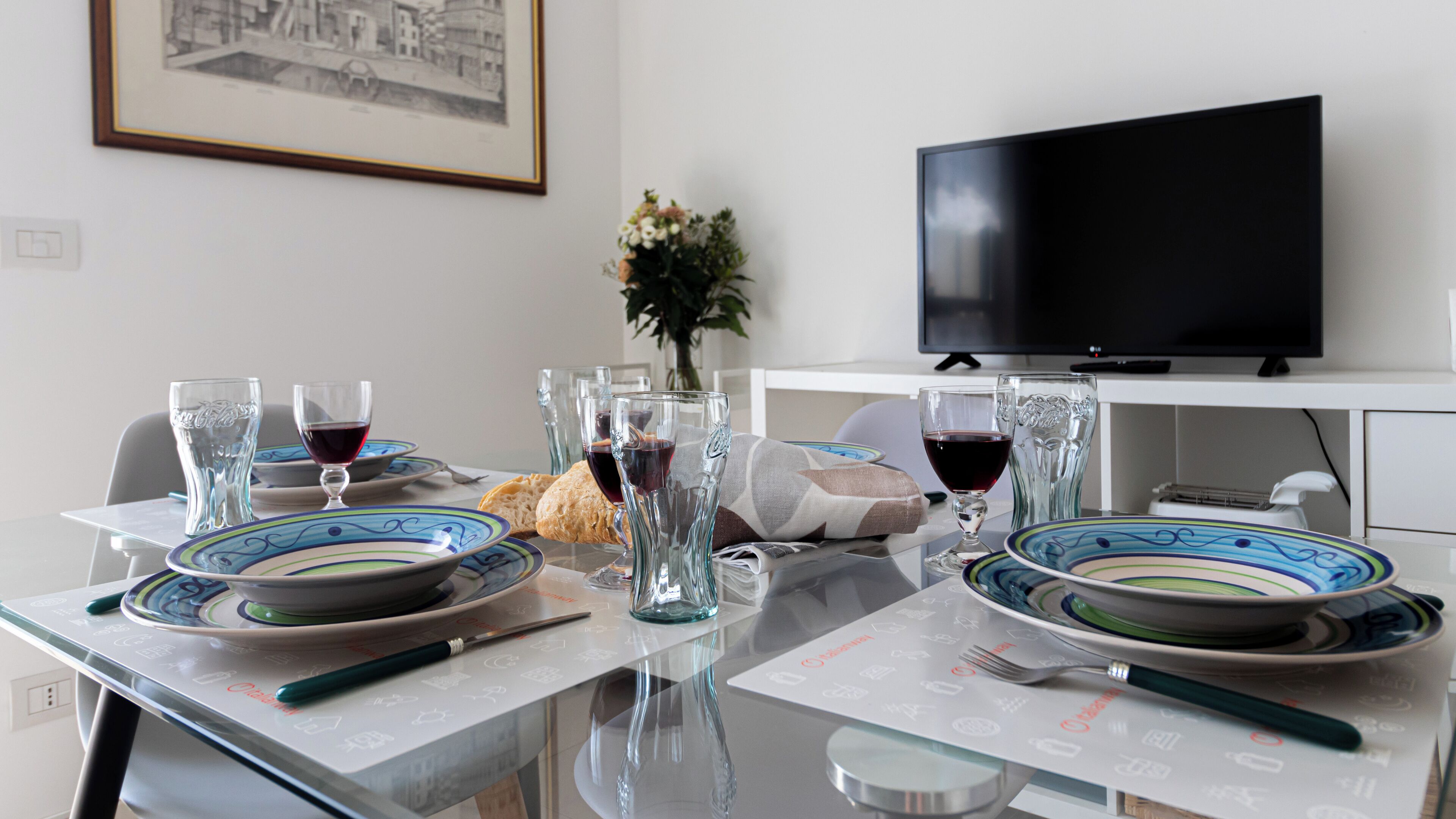 Een eettafel gedekt met borden, glazen en bestek, een vaas met bloemen en een televisie op een standaard.