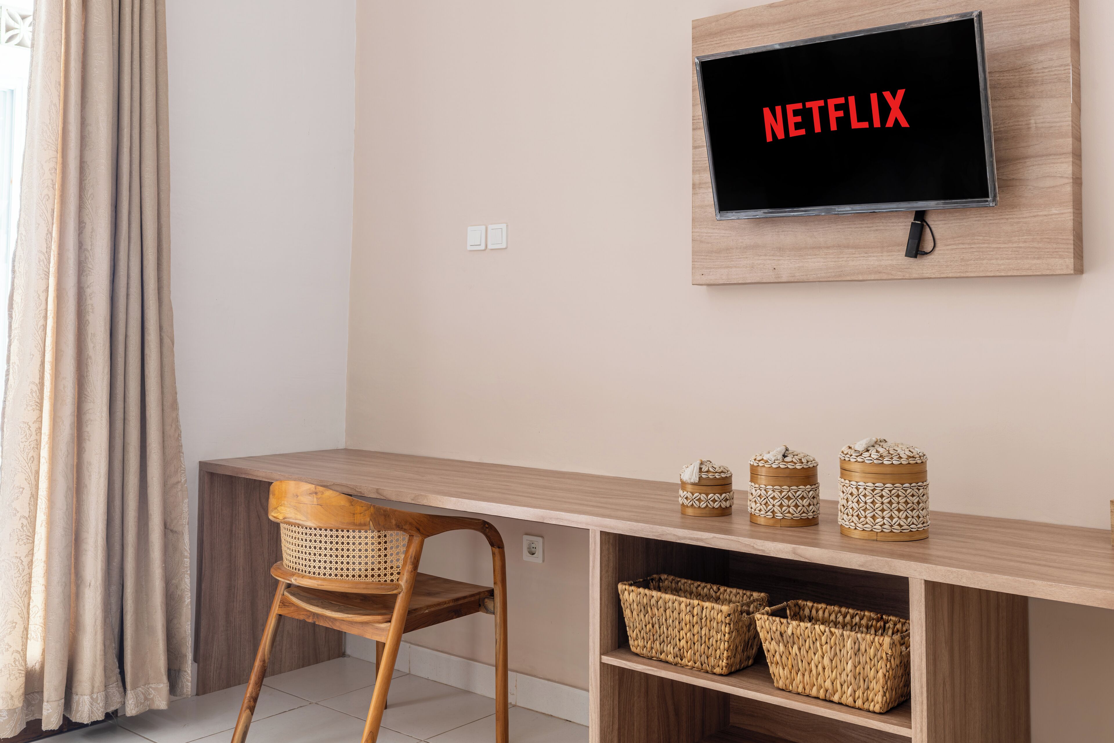 A room with a wooden desk, a chair, a TV mounted on the wall displaying "NETFLIX", and wicker baskets on the desk.