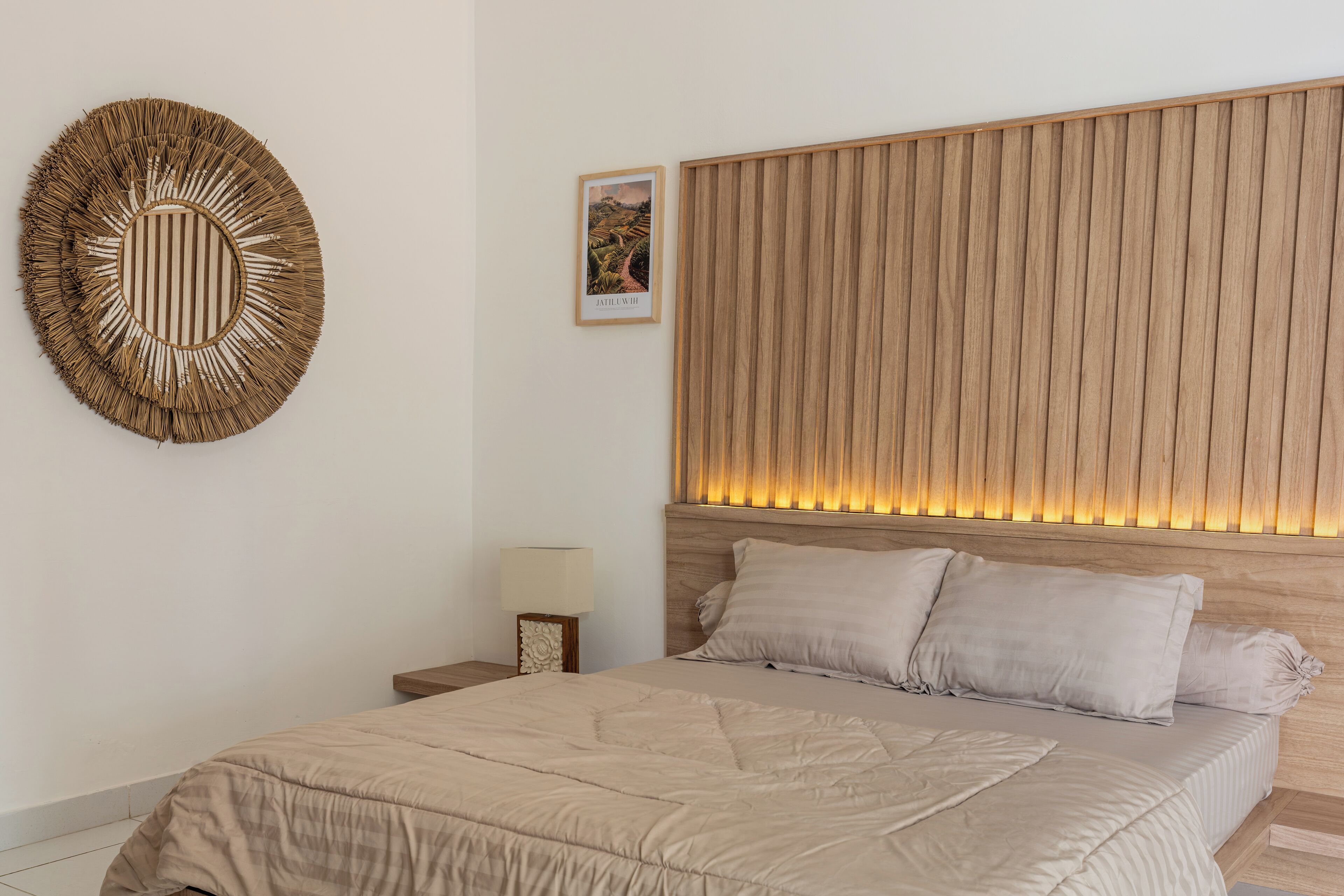 A bedroom with a large bed, a headboard with vertical wooden slats, a wall-mounted circular mirror, and a small bedside table with a lamp.