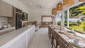 Une cuisine moderne dotée d’un îlot central, d’un coin repas avec des chaises en osier et de grandes fenêtres offrant une vue sur le jardin.