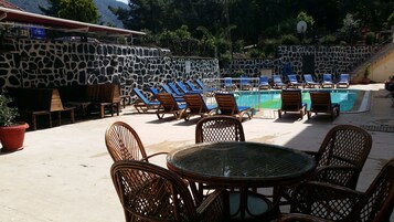 Área de piscina com espreguiçadeiras e uma mesa, cercada por paredes de pedra e plantas em vasos.
