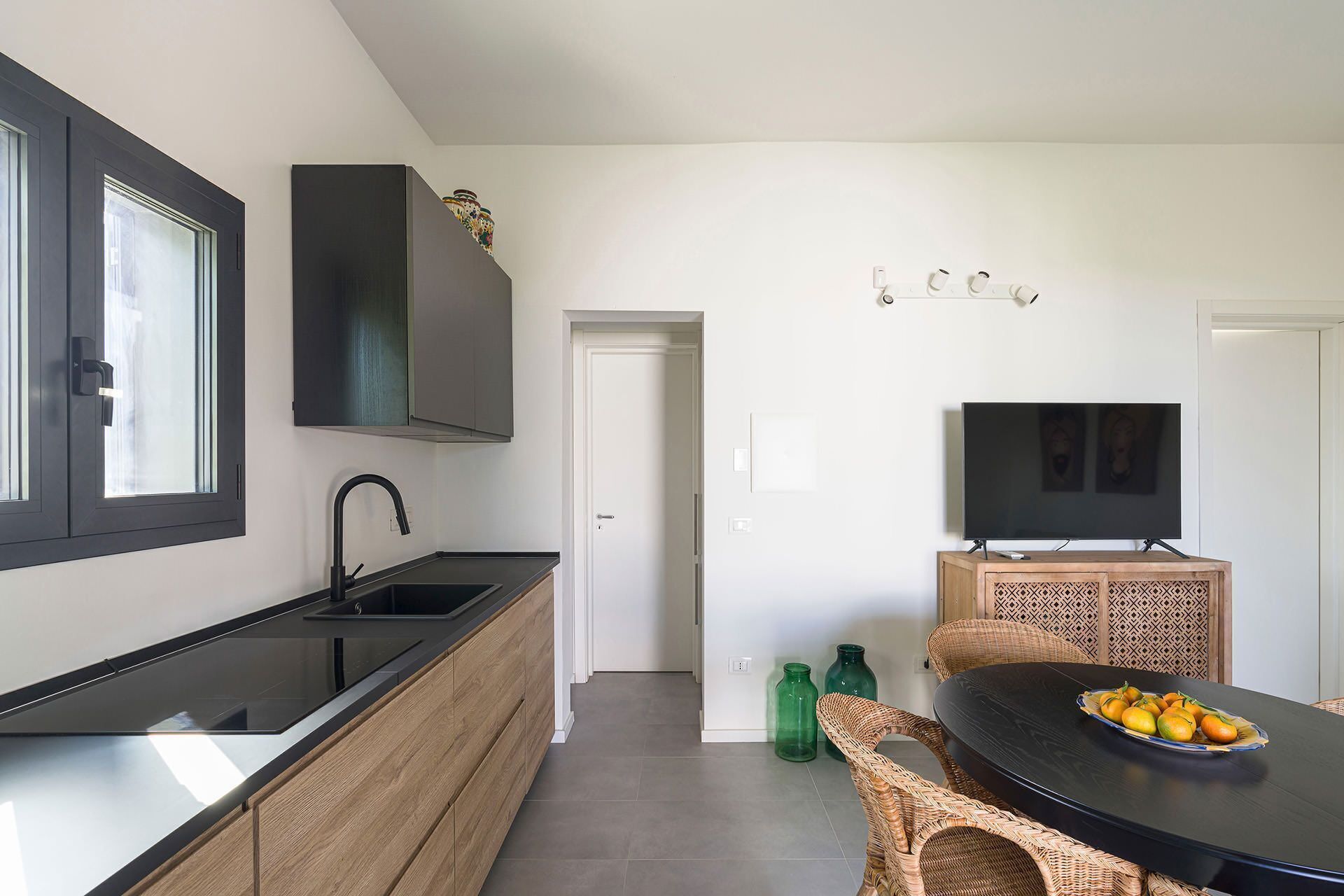 Una cocina moderna con encimera negra, pileta integrada y un mueble de madera. Hay un comedor con una mesa negra y sillas de mimbre, y un televisor montado en la pared.