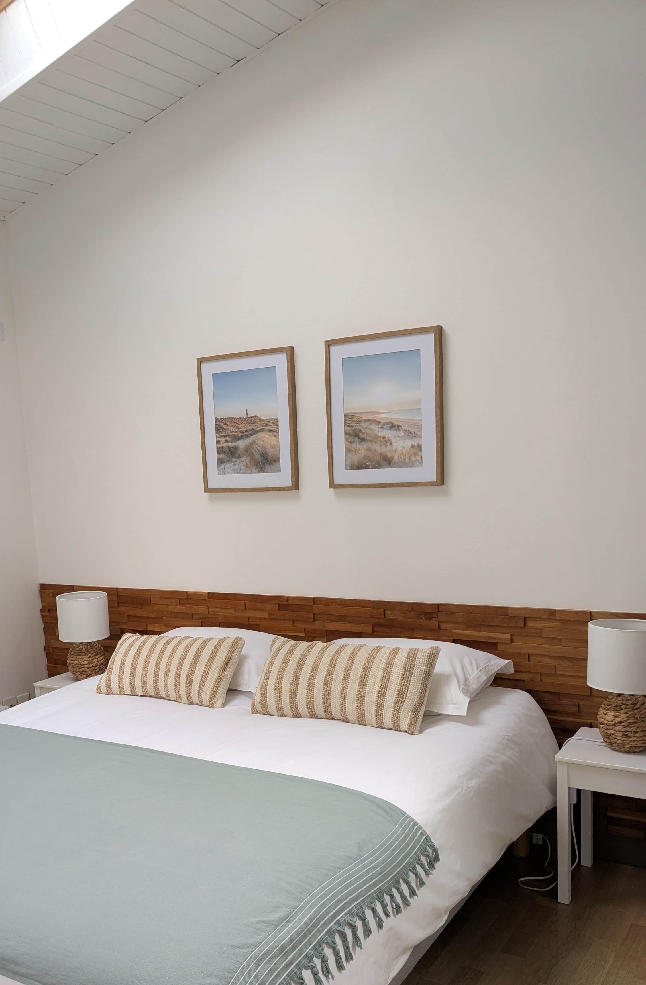 Une chambre à coucher avec une tête de lit en bois, un lit agrémenté d’oreillers à rayures, deux tableaux représentant des paysages encadrés au mur et une table de chevet avec une lampe.