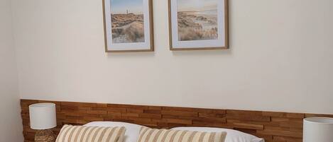Une chambre à coucher avec une tête de lit en bois, un lit agrémenté d’oreillers à rayures, deux tableaux représentant des paysages encadrés au mur et une table de chevet avec une lampe.