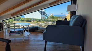 A cozy loft bedroom with a sloped ceiling, a comfortable blue sofa, a small round table, and a view of the sea and houses. - L'Abri de la Plage, L'Abri de la plage - Maison de bord de mer en Bretagne (Lampaul-Plouarzel)